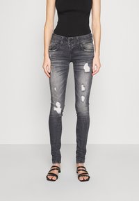 Monosuit negro con jeans ajustados grises que presentan desvanecidos, parches desgastados, cinco bolsillos y cierre de botón. La modelo lleva sandalias negras de tiras.