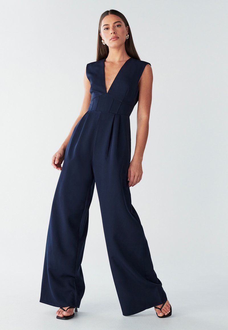 Temno modr kombinezon z globokim V-izrezom, kratkimi rokavi in širokimi hlačnicami. Ima oprijeten pas in gladko teksturo, podobno satenasti tkanini.