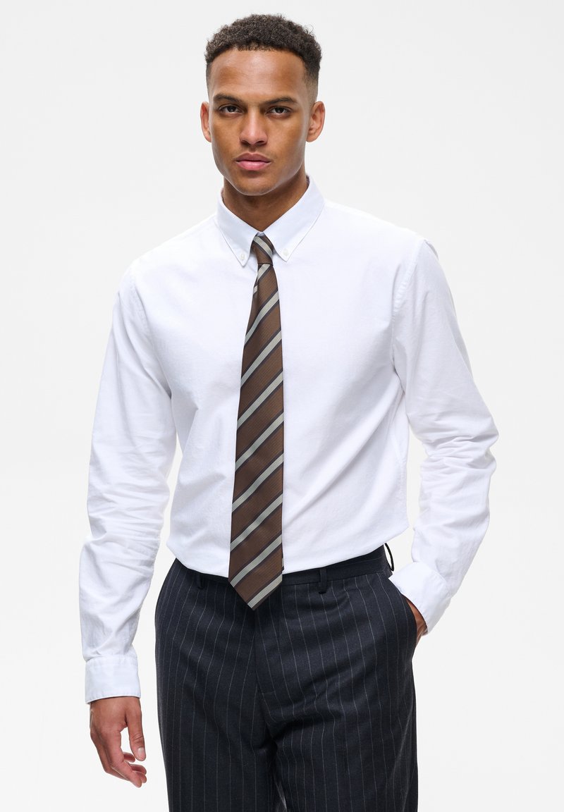 Homme portant une chemise blanche boutonnée, une cravate rayée marron et un pantalon sombre à fines rayures, une main dans la poche, sur un fond uni.