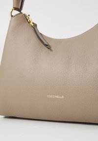 Borsa a mano in pelle beige con finitura testurizzata, forma curva e dettaglio del logo dorato. Presenta una tracolla corta e accenti di cuciture sottili.