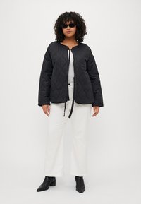 Zwarte gewatteerde jas met een ronde halslijn, sluiting aan de voorkant en lange mouwen. Gekleed met een wit top en een brede witte broek.