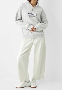 Grauer Fleece-Pullover mit halbem Reißverschluss und dem Schriftzug "MARSEILLE". Kombiniert mit weißen, weit geschnittenen Hosen und weißen Sneakern, zeigt er einen lässigen Stil.