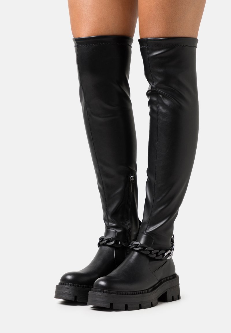 Botas negras de plataforma por encima de la rodilla con suelas gruesas y detalle decorativo de cadena, llevadas con piel visible sobre las botas.