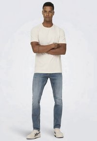 Beige bomulls t-shirt med kort ärm, kombinerad med ljusblå denimjeans. Modellen bär vita sneakers med marinblå accenter.