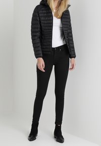 Frau in einer schwarzen Daunenjacke, weißem Shirt, schwarzen Skinny Jeans und schwarzen Ankle Boots, die vor einem schlichten hellen Hintergrund steht.