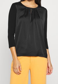 Svart topp med trekvartslånga ärmar, plisserad detalj vid halslinningen och slät textur, tillsammans med orange byxor.