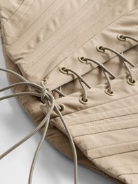 Beige stoffkorsett med vertikal søm, med øyer og snøring på den ene siden. Strukturert materiale med glatt overflate og en formet fasong.