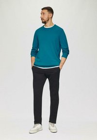 Türkiser Baumwoll-Sweatshirt mit Rundhalsausschnitt, über einem weißen Shirt getragen, kombiniert mit schwarzen Hosen und weißen Sneakers. Minimalistisches Design.