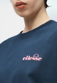 Άτομο που φοράει μπλε σκούρο φούτερ με στρογγυλή λαιμόκοψη και μικρό ροζ λογότυπο «ellesse» στο στήθος, με τα χείλη ορατά αλλά τα μάτια εκτός κάδρου.