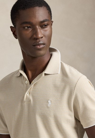 Μπεζ και λευκό ριγέ πόλο με γιακά, με μικρό κεντητό λογότυπο στο αριστερό στήθος και λεπτομέρεια υφής στο ύφασμα.