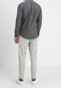 Chemise grise en coton à manches longues avec col boutonné et pli dans le dos, associée à un pantalon chino gris clair et des baskets blanches.