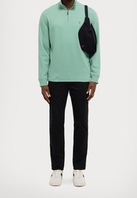 Mintgrön långärmad tröja med halv dragkedja och krage, svarta byxor och en svart axelväska. Skorna har flerfärgade accenter.