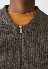 Brauner texturierter Pullover mit geripptem Stoff und einem metallischen Reißverschluss. Über einem senfgelben Shirt getragen, das ein rundes Ausschnittdesign zeigt.