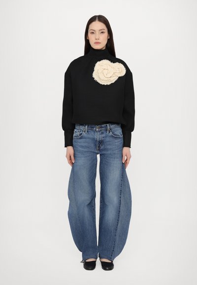 Maglione nero strutturato con collo alto e dettagli a fiori crema, abbinato a jeans in denim blu a gamba larga. Scarpe nere completano l'outfit.