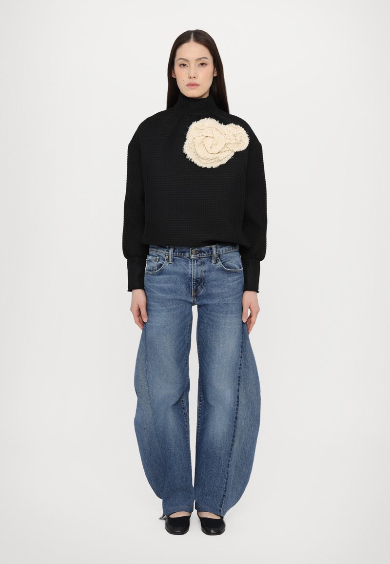 Sort tekstureret sweater med høj hals og cremefarvet blomsterdetalje, kombineret med brede blå denim jeans. Sorte sko fuldender outfittet.