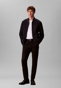 Veste aviateur marine portée sur un t-shirt blanc, pantalon noir et chaussures noires. Tissu lisse, coupe ajustée et design minimaliste.