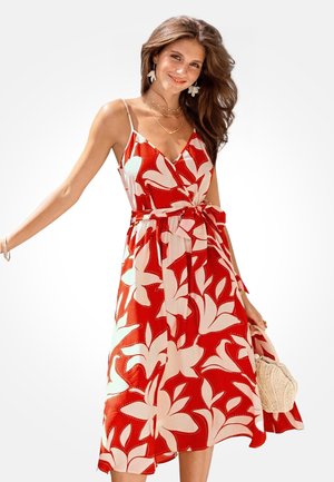 Woman wearing sleeveless red dress with large white floral pattern, holding woven handbag, smiling with long flowing hair.