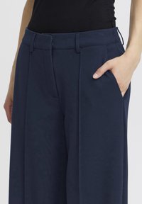 Prim-plan cu o persoană purtând pantaloni largi bleumarin cu talie înaltă, având mâna în buzunarul din dreapta, asortați cu un top negru.