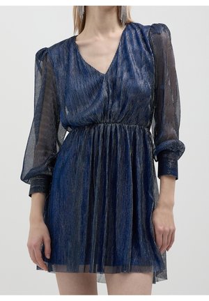 Femme portant une robe plissée métallique bleue avec des manches longues transparentes et un col en V, debout devant un fond clair uni.