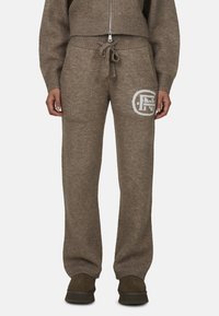 Brązowe dzianinowe spodnie dresowe z regulowanym paskiem w talii i białym logo. Posiadają ściągacze przy nogawkach oraz miękką teksturę.