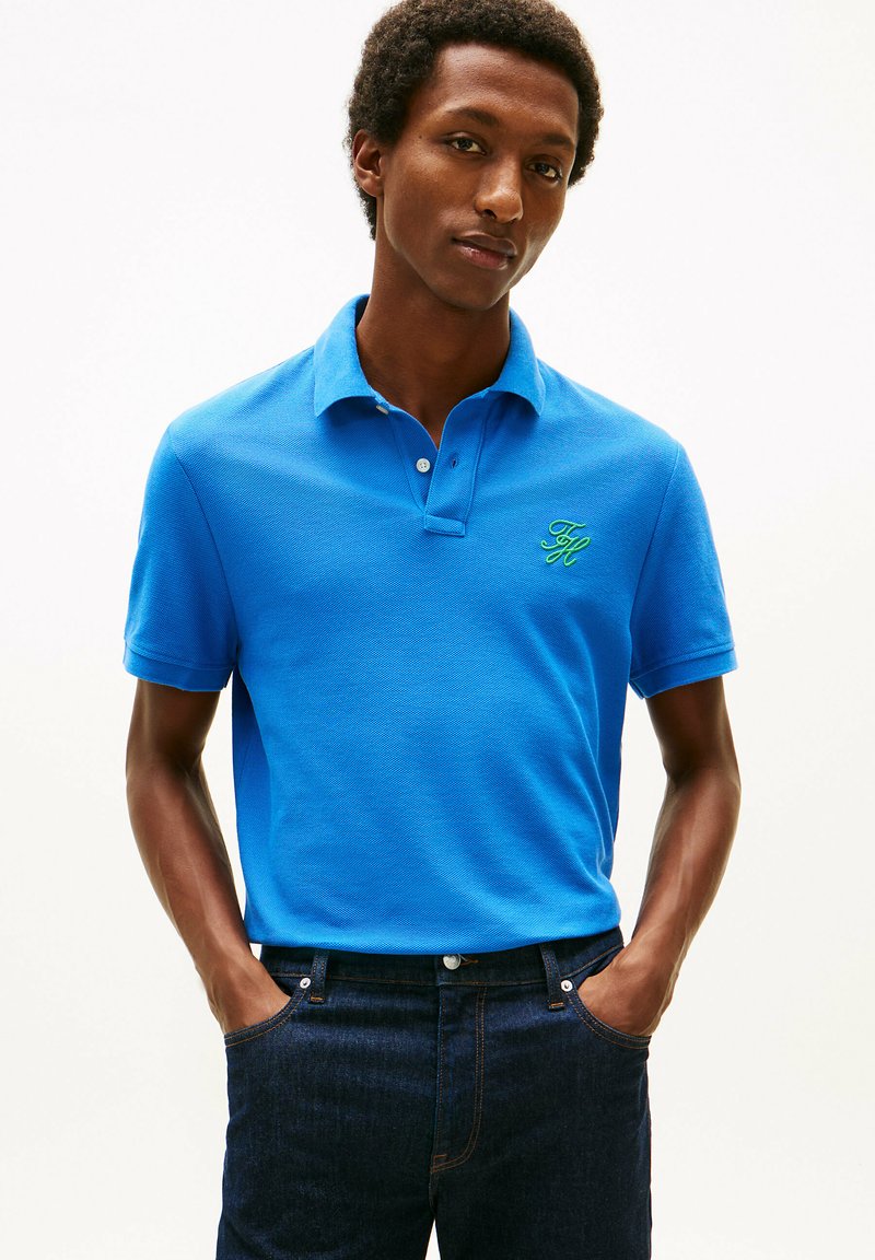 Giovane uomo che indossa una polo blu brillante con logo verde sul petto e jeans blu scuro, in piedi con le mani nelle tasche contro uno sfondo bianco.