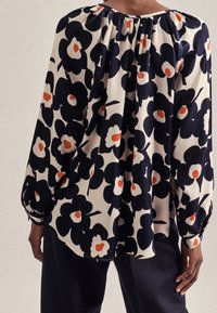 Blommig blus med ett mönster i marinblått, krämfärgat och orange. Har en rynkad halsringning och långa ärmar, tillverkad av ett mjukt tyg.