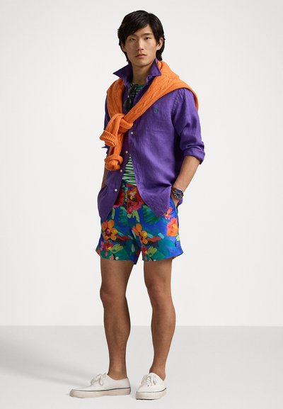 Modell trägt ein lila Leinenhemd, einen orangefarbenen Strickpullover um den Hals gebunden, ein gestreiftes grünes Hemd, floral gemusterte blaue Shorts und weiße Sneakers.