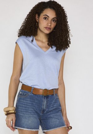 Donna con capelli ricci che indossa una canotta azzurro chiaro, pantaloncini di jeans, cintura marrone e braccialetti impilati al polso.