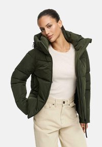Femme portant une doudoune olive sur un haut beige à côtes et un pantalon taille haute beige clair, posant avec une main dans la poche.