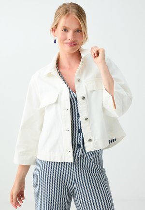 Smiling woman with blonde hair wears white buttoned jacket over blue and white patterned top and pants, holding jacket collar.