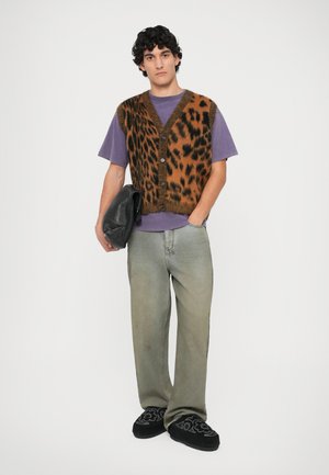 Kamizelka w wzór panterki nałożona na fioletowy T-shirt, połączona z wyblakłymi zielonymi dżinsami i czarnymi haftowanymi butami slip-on, trzymająca czarną torbę.