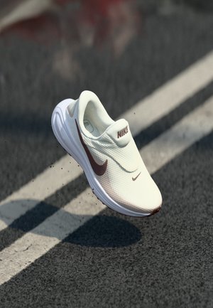 Chaussure de course Nike blanche slip-on avec logo swoosh marron flottant au-dessus d'une route asphaltée avec des marquages blancs.