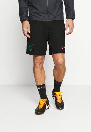 Homme portant un short de sport noir avec le logo du FC Barcelone, des chaussettes Nike noires et des crampons noirs avec des lacets orange vif.