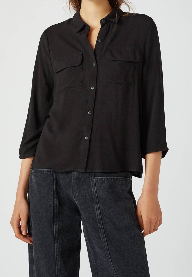 Femme portant une chemise noire boutonnée avec des poches à rabat sur le devant et manches trois-quarts, associée à un jean large en denim gris foncé.