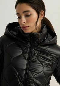 Czarna, pikowana kurtka puchowa z wysokim kołnierzem i kapturem. Posiada gładką, błyszczącą fakturę oraz zamek błyskawiczny z przodu. Charakteryzuje się wyrazistym, zakrzywionym wzorem szwów.