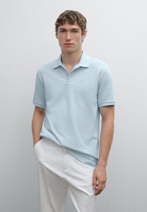 Polo shirt azzurro chiaro in morbido cotone con colletto, maniche corte e pattina con tre bottoni. Indossato con pantaloni bianchi. Stile minimalista.