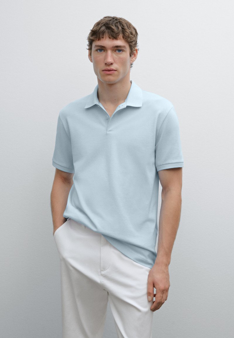 Light blue polo shirt in soft cotton with a collar, short sleeves, and a three-button placket. Worn with white pants. Minimalist style.