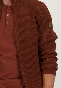 Bruine geribbelde gebreide cardigan met een hoge kraag, voorzien van een rits en smalle manchetten, gedragen over een donker kastanjebruine shirt met drie knopen.