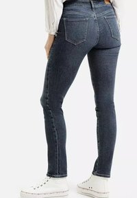 Osoba ubrana w ciemnoniebieskie dopasowane dżinsy, białą koszulę i białe wysokie trampki, pokazana od tyłu na jednolitym tle.