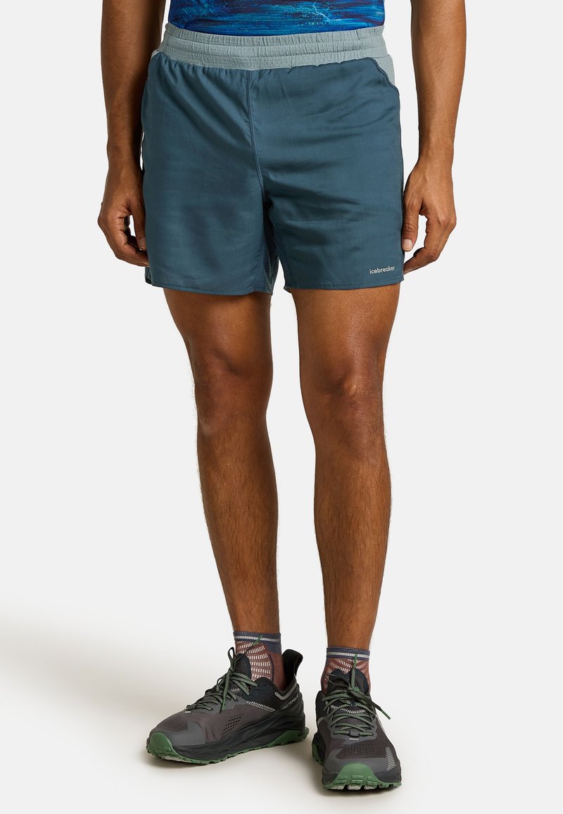 Men's athletic shorts in dark teal, featuring an elastic waistband and lightweight fabric. Paired with gray and green running shoes.