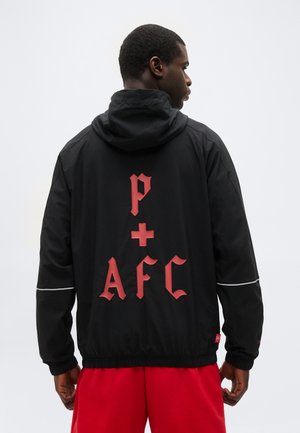 Uomo con giacca nera con cappuccio, con lettere rosse in stile gotico "P + AFC" sul retro, abbinata a pantaloncini rossi, di spalle su sfondo neutro.