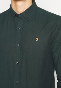 Camicia button-up verde scuro in un tessuto morbido, con colletto a punta, bottoni neri e un piccolo logo giallo ricamato sul petto.