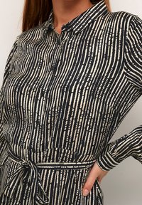 Camicia a motivi nera e crema con righe verticali, design con bottoni, colletto e cintura in vita. Presenta un tessuto testurizzato e maniche lunghe.