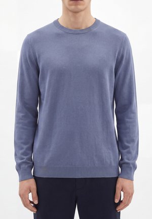Hombre que lleva un jersey ajustado de manga larga y cuello redondo en azul apagado con un sutil logo "AM" en el pecho izquierdo, combinado con pantalones oscuros.