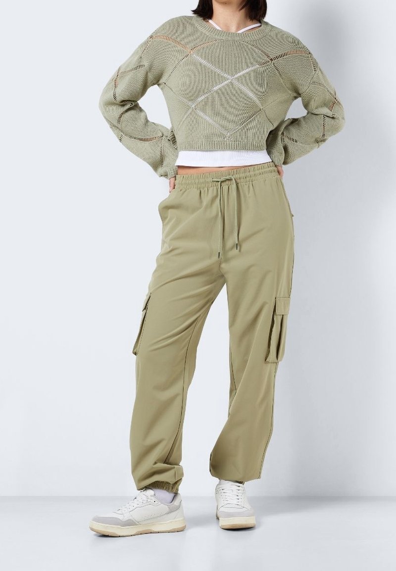 Garnk sweater i ljusgrön med diamantmönster, tillsammans med beige cargobyxor med fickor, elastisk midjeband och muddar vid anklarna. Vita sneakers.