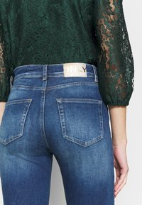 Jeans de ganga azul skinnies com uma cintura alta, apresentando dois bolsos traseiros e uma etiqueta de marca em dourado na cintura.