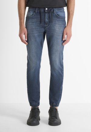 Hombre con vaqueros jogger azules con puños elásticos y zapatillas negras, de pie frente a un fondo blanco liso.