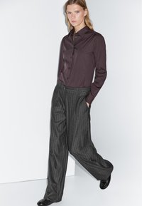 Vrouw die met haar handen in de zakken loopt, gekleed in een donkerbruine button-up shirt en grijs gestreepte wijde broek met zwarte schoenen.