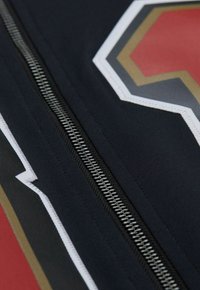 Schwarze Jacke mit einem zentralen Reißverschluss und mutigen geometrischen Mustern in Rot, Weiß und Gold. Texturiertes Gewebe mit glatten Akzenten.
