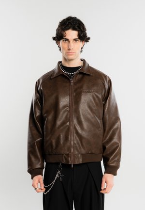 Jeune homme aux cheveux courts, bouclés et foncés, portant une veste en cuir marron zippée sur un pantalon noir avec un accessoire chaîne argentée.